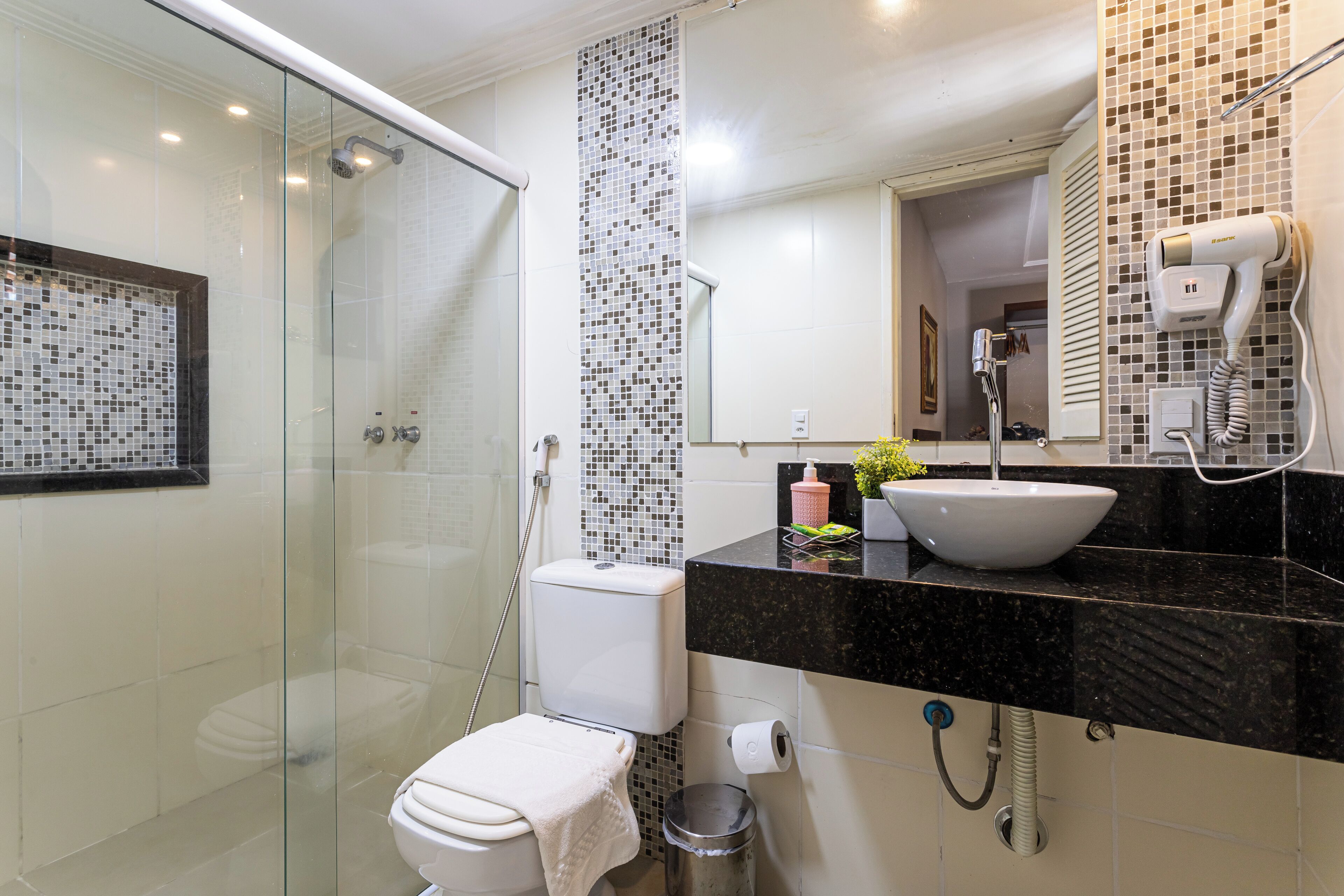 A modern bathroom with a glass-enclosed shower, a white toilet, a black countertop with a sink, and a hairdryer mounted on the wall.