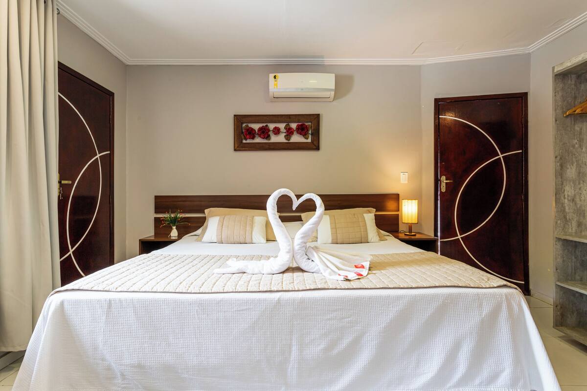 Une chambre d’hôtel avec un lit, un climatiseur fixé au mur, un tableau encadré et une table de chevet avec une lampe.