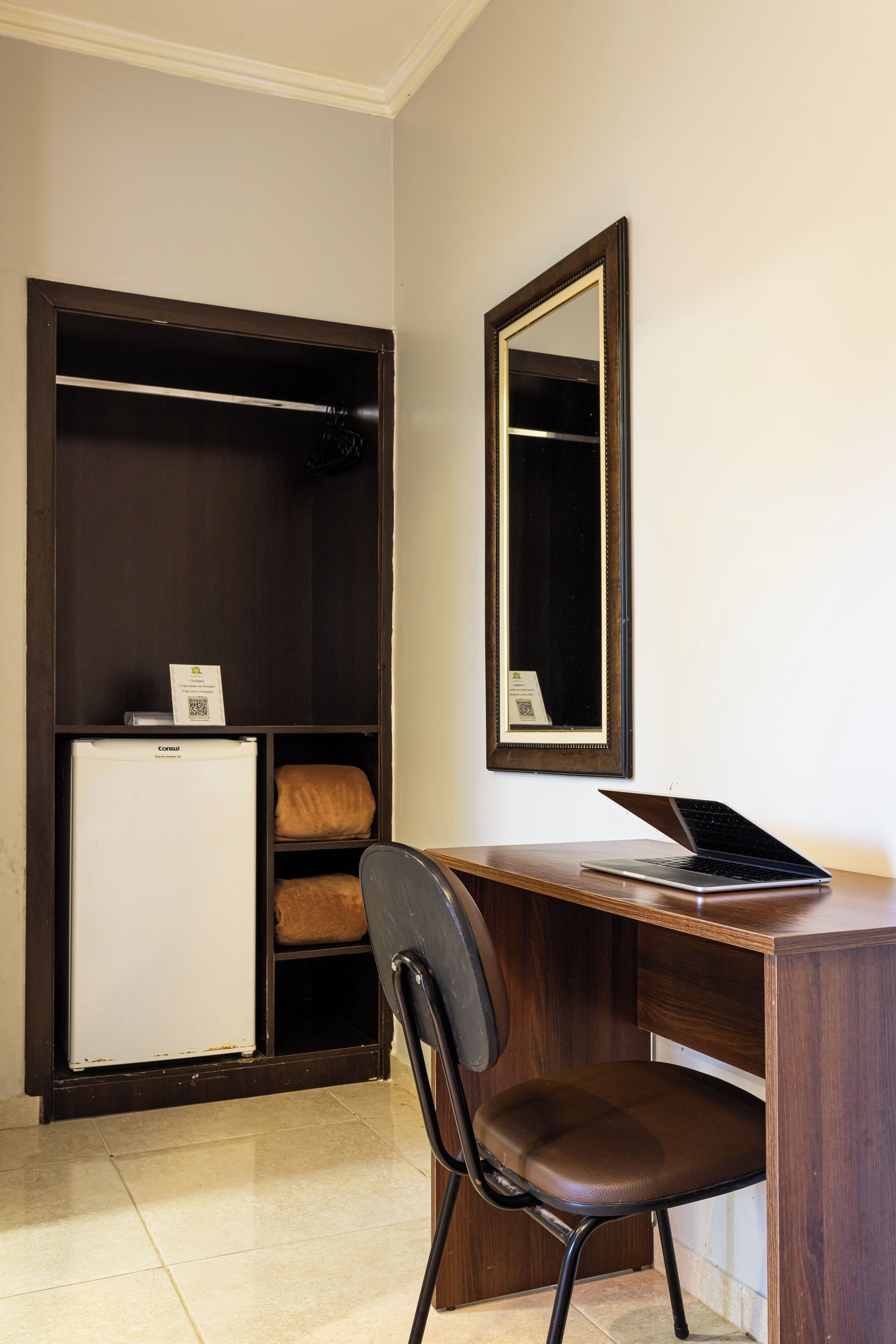 A room with a desk, chair, wardrobe, and a small white refrigerator.