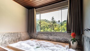 A bathroom with a marble countertop, a bathtub filled with bubbles, a window with a view of greenery, and a potted plant. - Pousada Recanto de Moriá (Itatiaia)