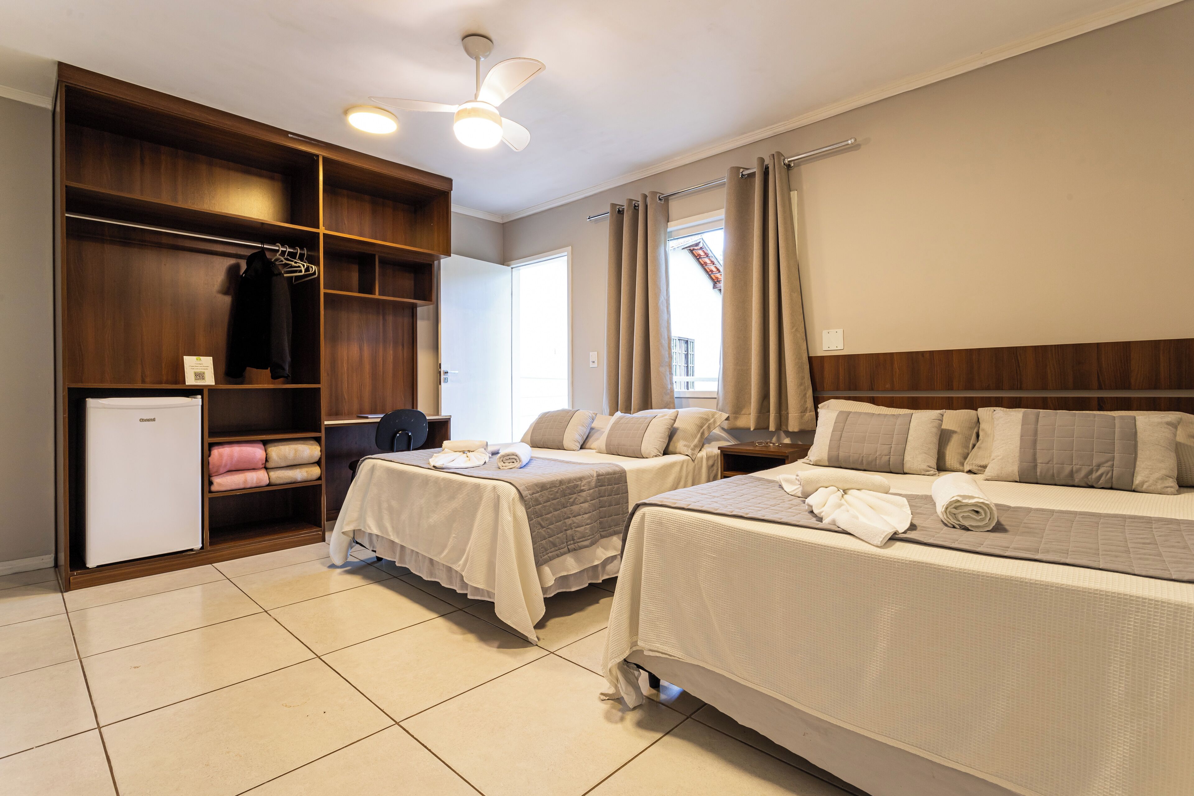 A hotel room with two beds, a wardrobe, a small fridge, and a ceiling fan.
