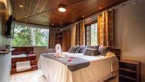 A bedroom with a large bed, a wooden ceiling, a TV mounted on the wall, and a window with curtains. - Pousada Recanto de Moriá (Itatiaia)