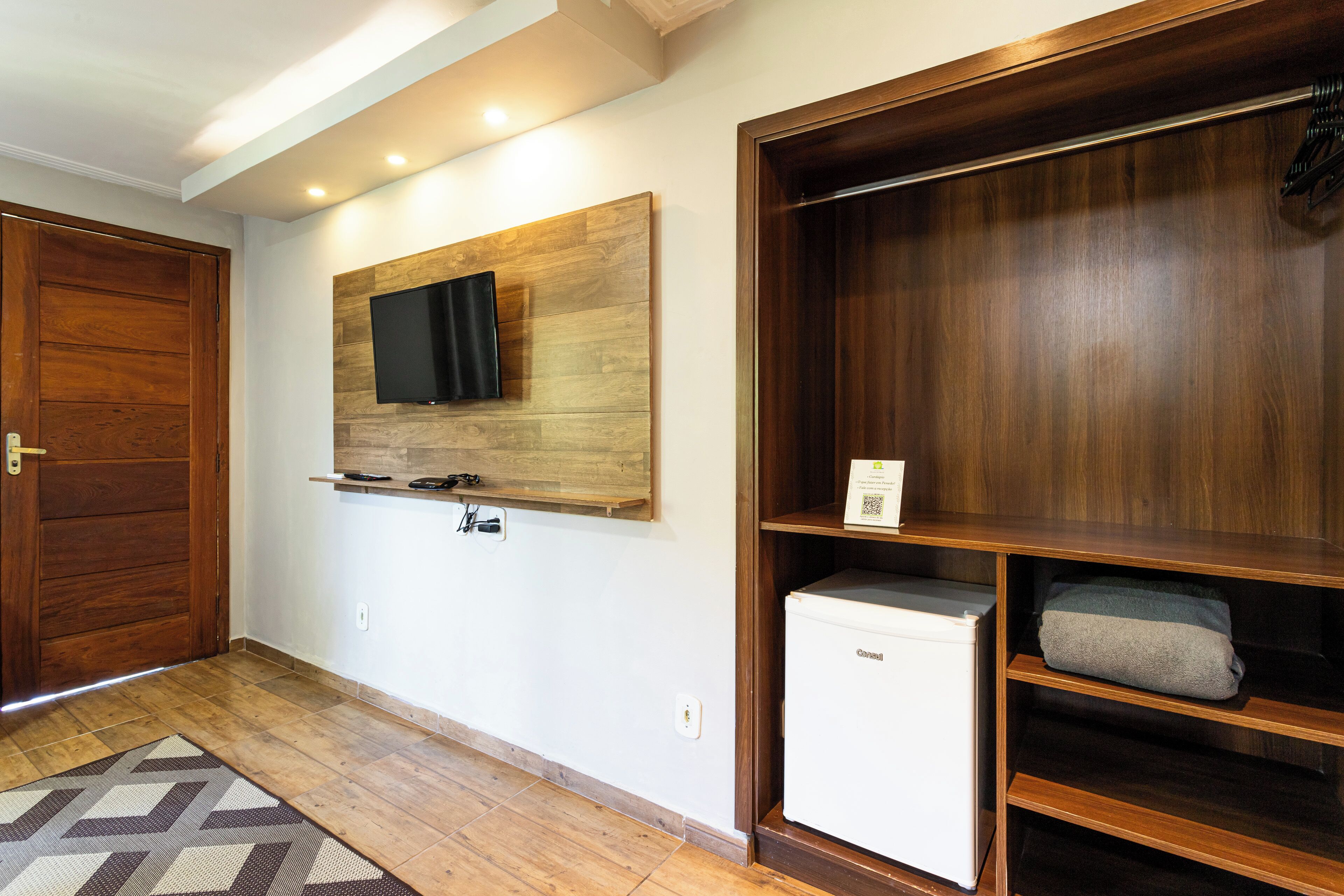 A hallway with a wooden door, a mounted television, a small refrigerator, and a wooden shelving unit.