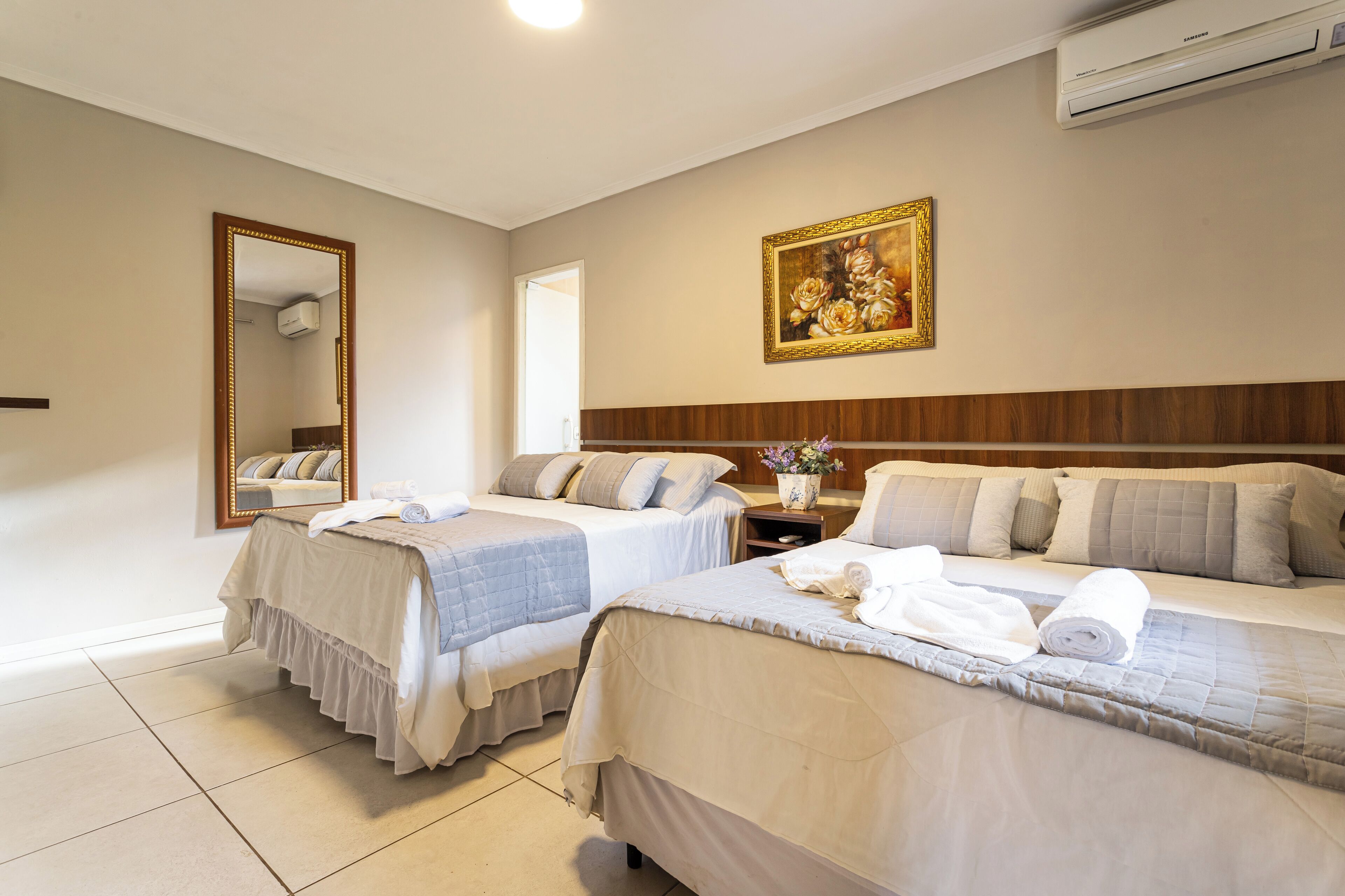 A hotel room with two beds, a large mirror, a painting, and an air conditioning unit.