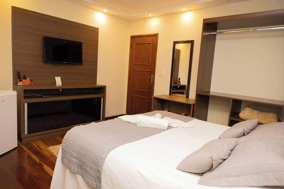 Une chambre d’hôtel avec un lit, un téléviseur fixé au mur, une porte en bois, un miroir et un petit bureau avec une lampe.