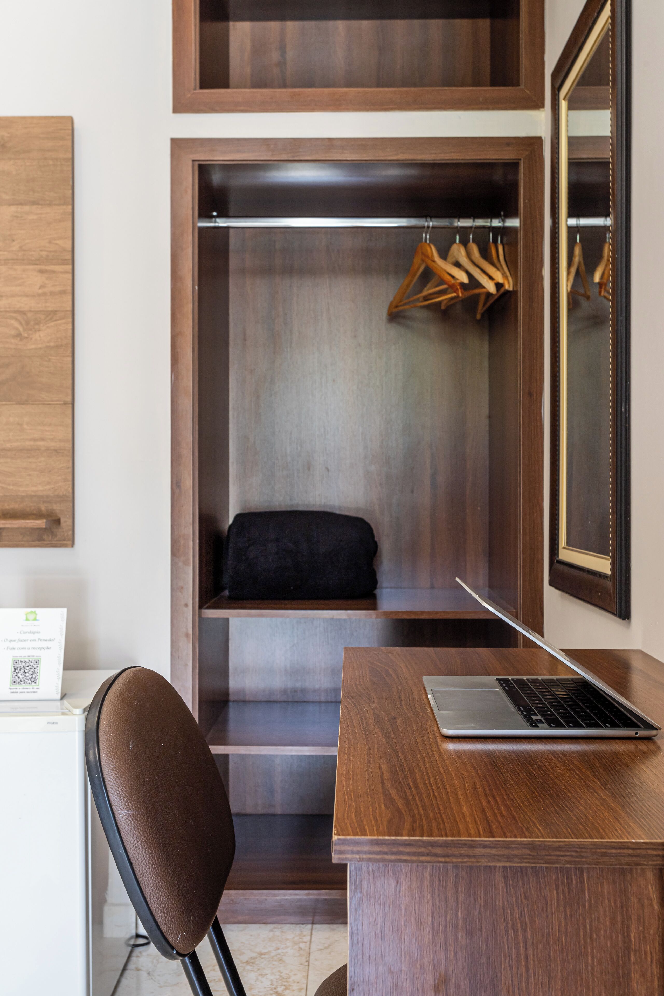 A wooden desk with a laptop, a chair, a coat rack with hangers, a black cushion, and a mirror on the wall.
