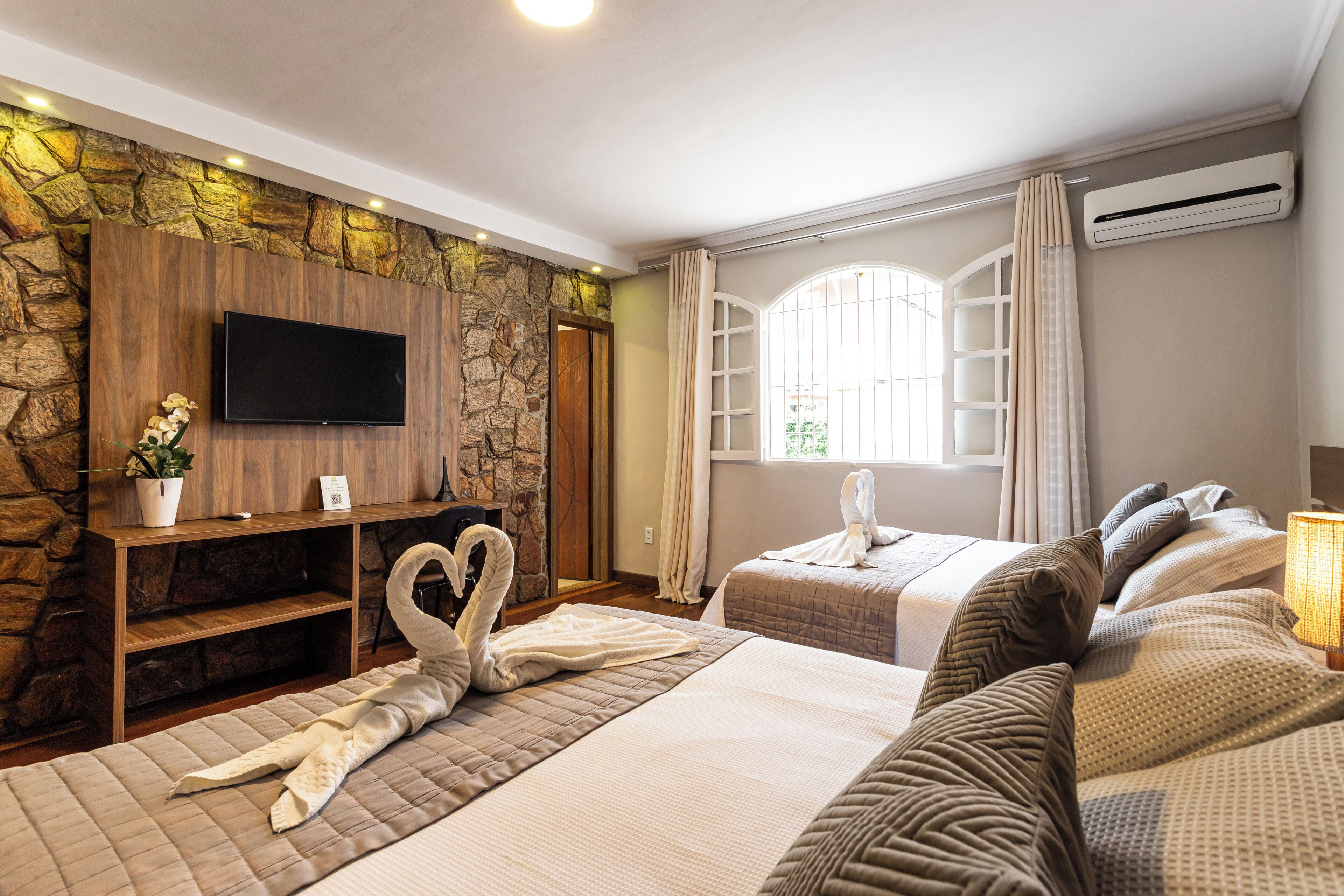 A hotel room with a stone wall, a flat-screen TV, two beds with heart-shaped towels, a desk with a plant, and a window with curtains.
