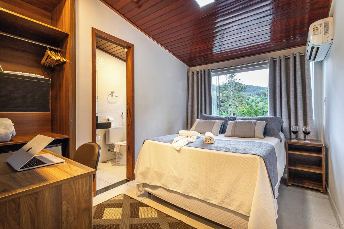 Une chambre à coucher comprenant un lit, un bureau avec un ordinateur portable, un espace douche et une fenêtre avec des rideaux.