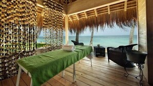Une terrasse en bois avec une table de massage, des chaises et une structure à toit de chaume offrant une vue sur l’océan.