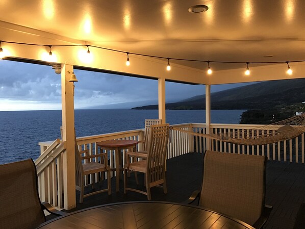 A covered outdoor seating area with wooden furniture and string lights, overlooking a body of water and a distant coastline. - RELAXING, Away-from-it-All home on the water with small Natural Pool (Milolii)