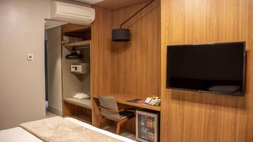 Una habitación pequeña con una cama, un escritorio con silla, un televisor en la pared y un refrigerador compacto.