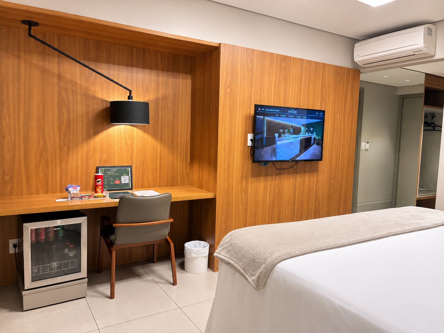 A hotel room with a bed, a desk with a computer, a television mounted on the wall, and an air conditioning unit.