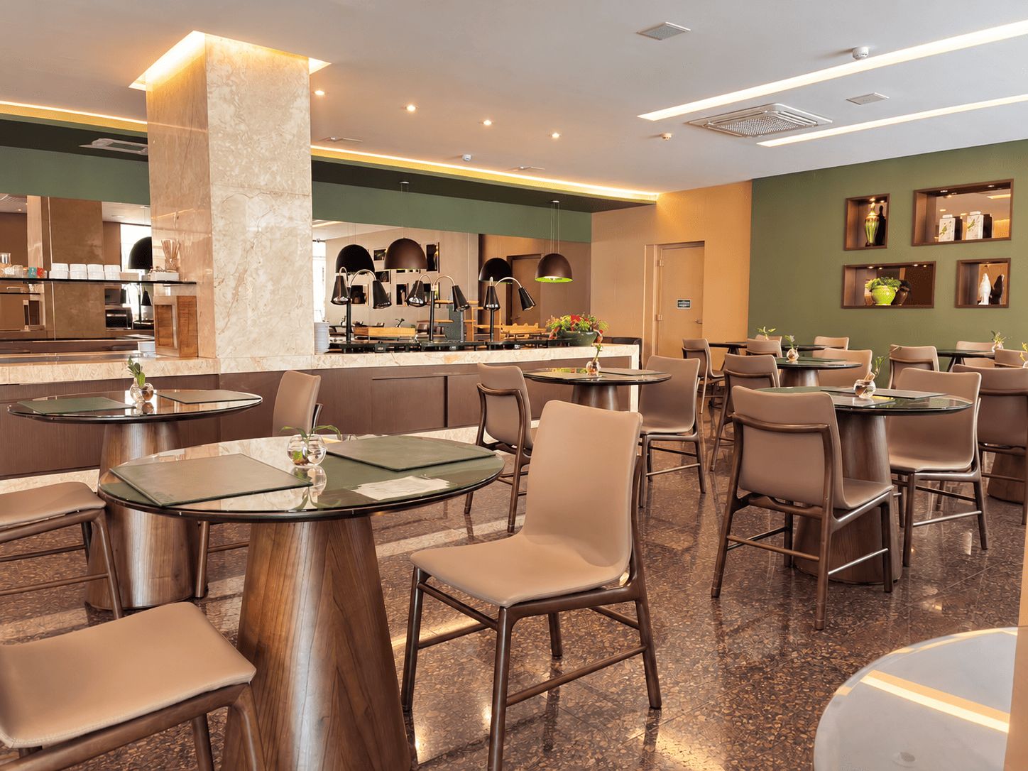 Ein modernes Restaurant mit runden Tischen und Stühlen, einem sichtbaren Küchenbereich und einer grünen Akzentwand.