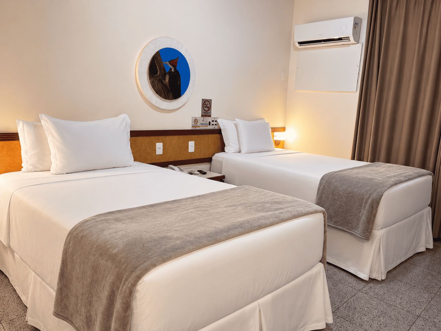 Ein Hotelzimmer mit zwei Betten, einer Wand-Klimaanlage, einem runden Spiegel und einem kleinen Tisch mit Telefon und Fernbedienung.