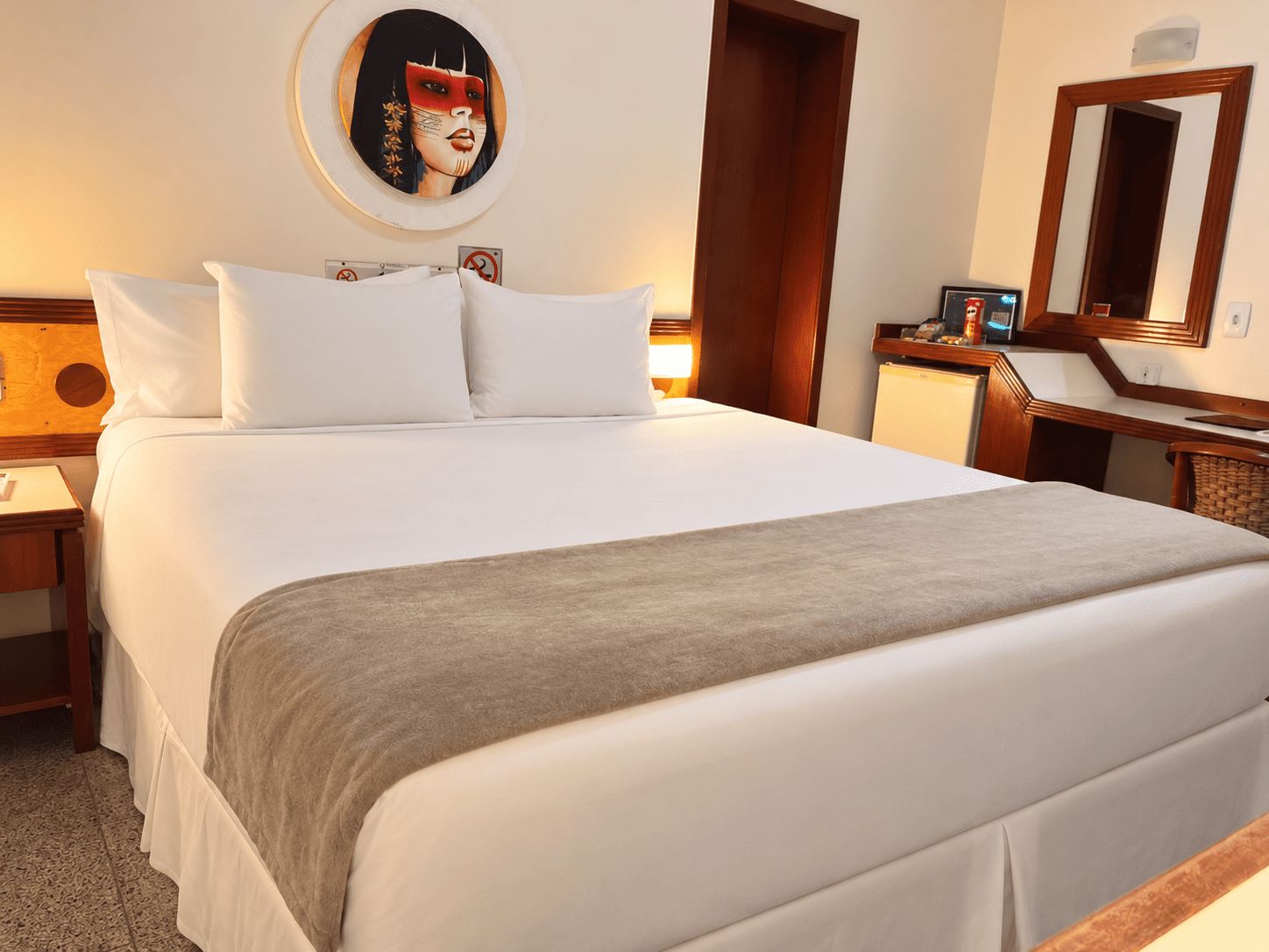 A hotel room with a large bed, a round wall clock, a wooden desk, and a mirror.