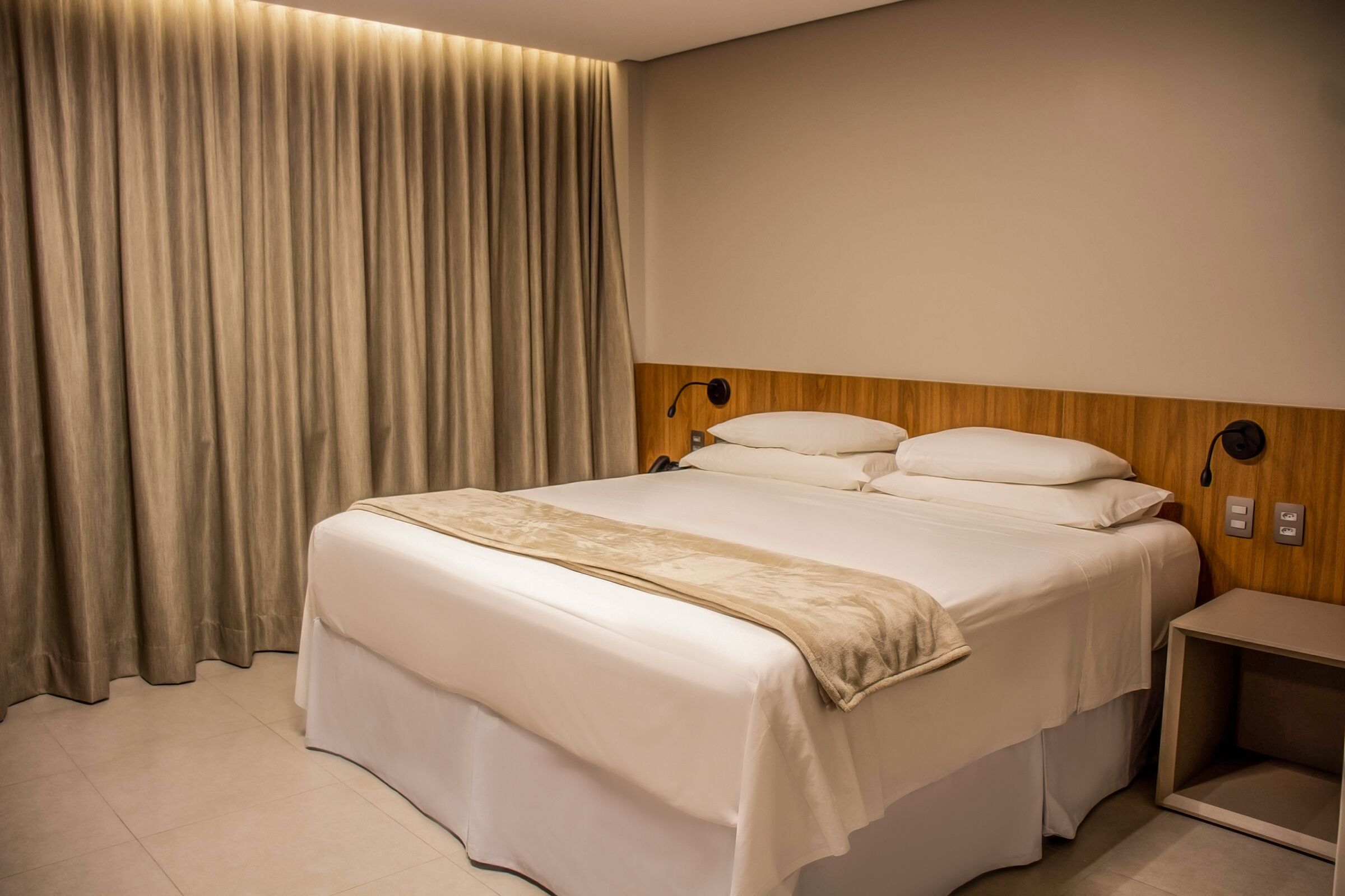 A hotel room with a large bed, a wooden headboard, a bedside table, and a curtain.
