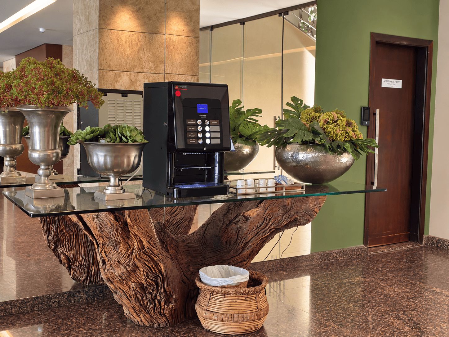 A coffee machine on a glass table with a wooden tree trunk base, surrounded by decorative pots and a wicker basket.
