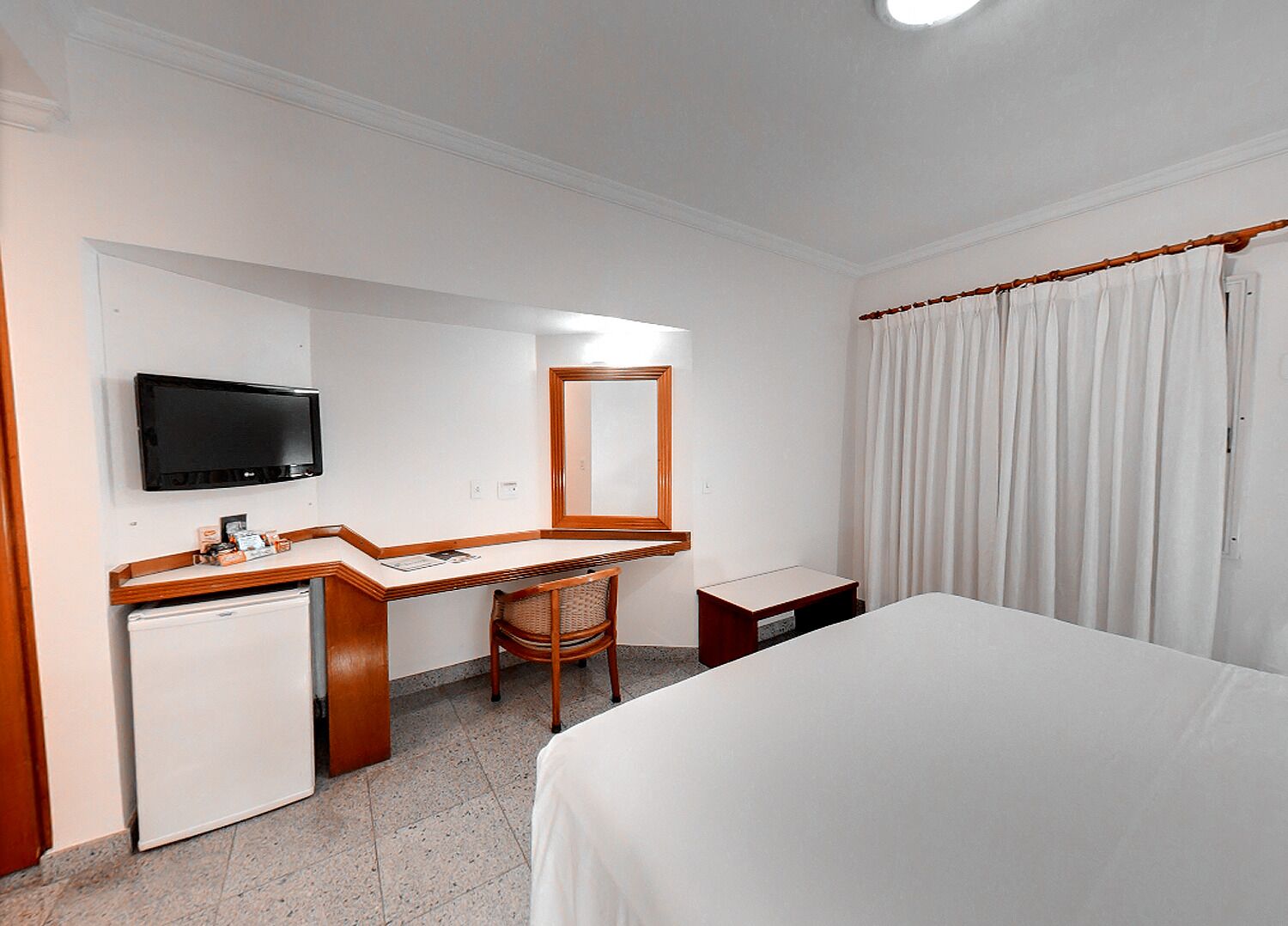 Quarto de hotel com cama, escrivaninha com televisão, pequeno frigorífico, cadeira e janela grande com cortinas.