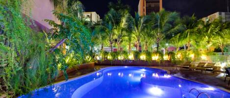 A swimming pool with a curved edge, surrounded by palm trees and plants, with a building in the background.