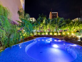 Una piscina de borde curvo, rodeada de palmeras y plantas, con un edificio al fondo.