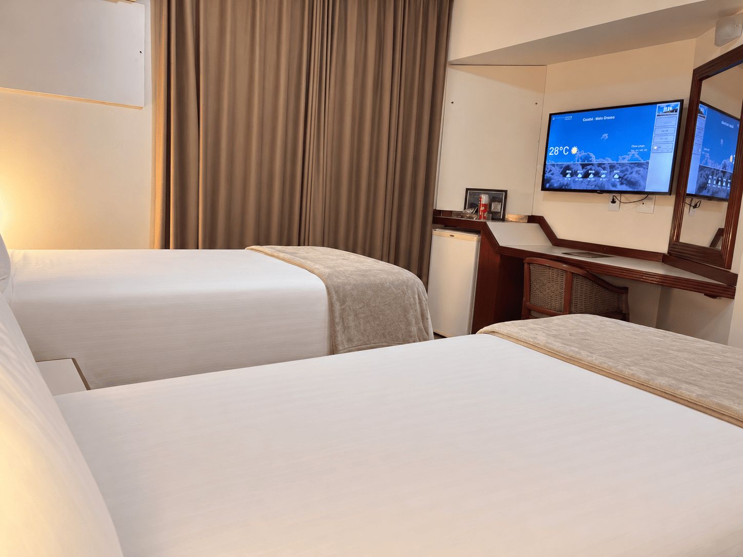 A hotel room with two beds, a television mounted on the wall, and a small desk with a mirror.