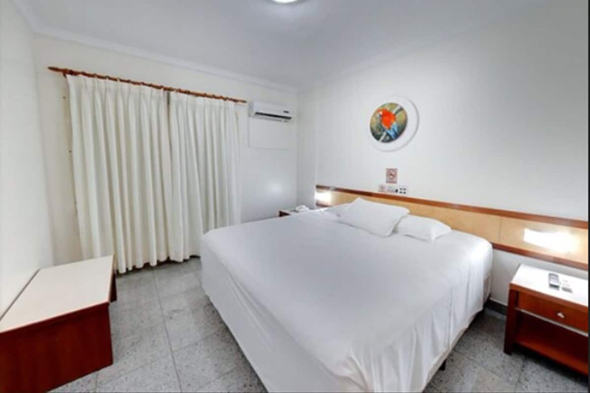 Une chambre d’hôtel avec un grand lit, une table de chevet, une commode, un climatiseur fixé au mur et un tableau accroché au mur.