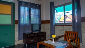 A room with a television on a wooden cabinet, a chair, a table with a vase of flowers, and a window with curtains. - Sabina Guesthouse (Kamala)