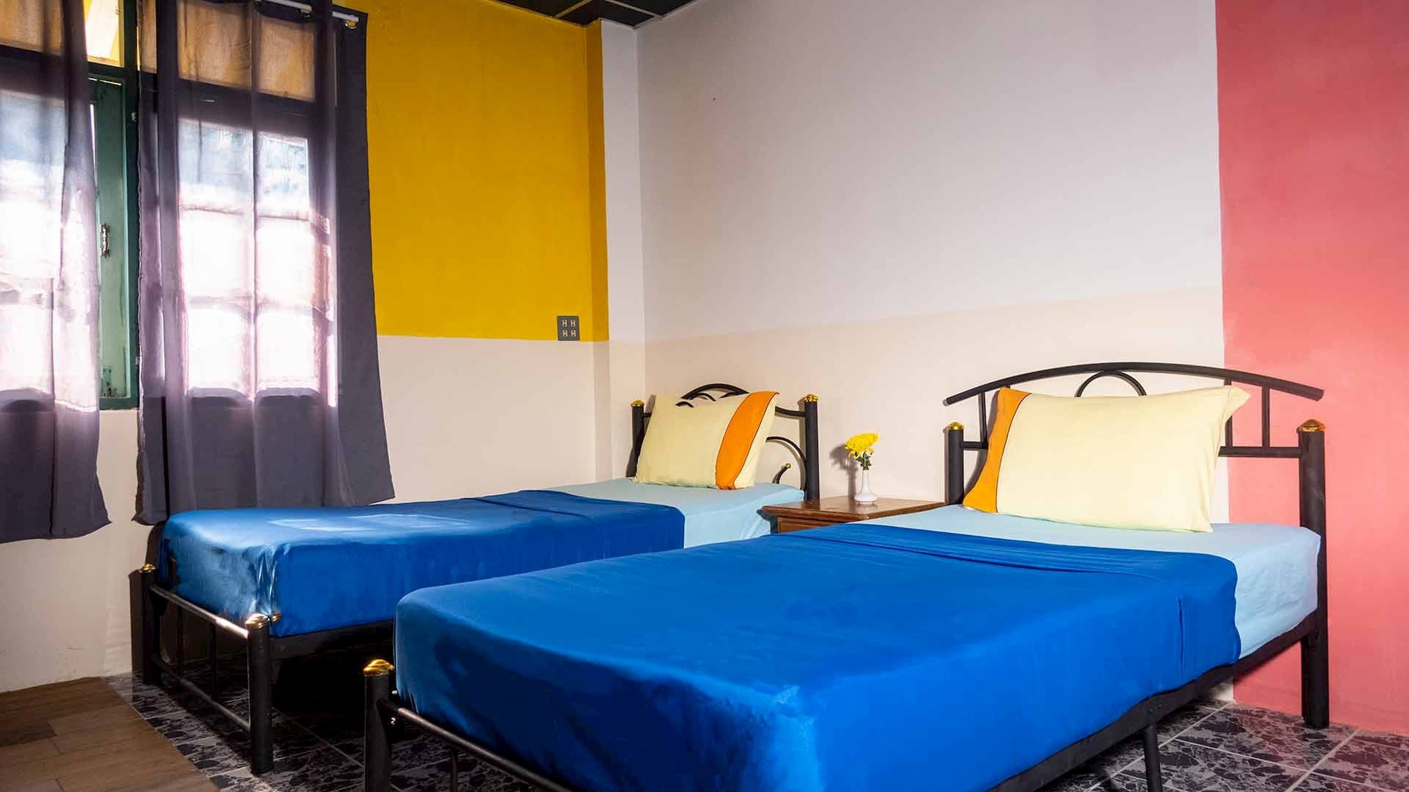 A room with two beds, each with blue bedding and yellow pillows, a window with sheer curtains, and a yellow wall.