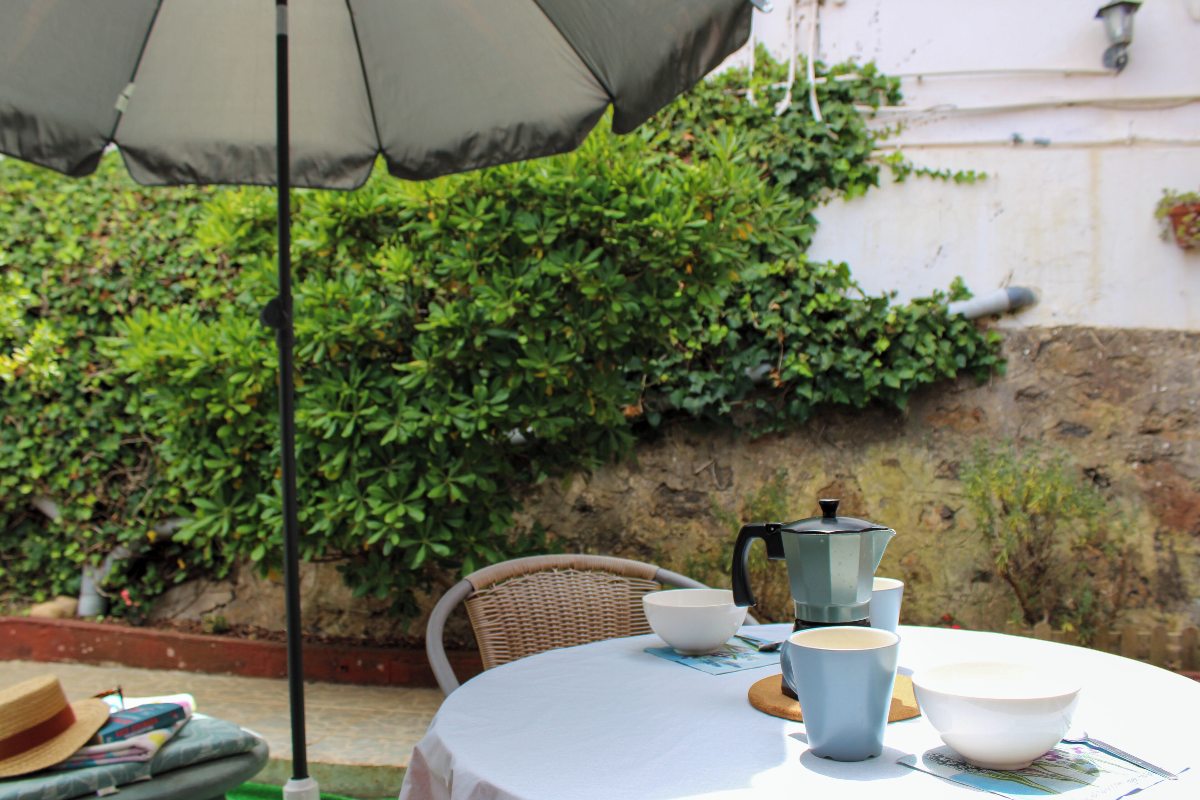 An outdoor table with a white tablecloth, a wicker chair, a coffee pot, and a hat on the table.