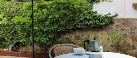 An outdoor table with a white tablecloth, a wicker chair, a coffee pot, and a hat on the table.