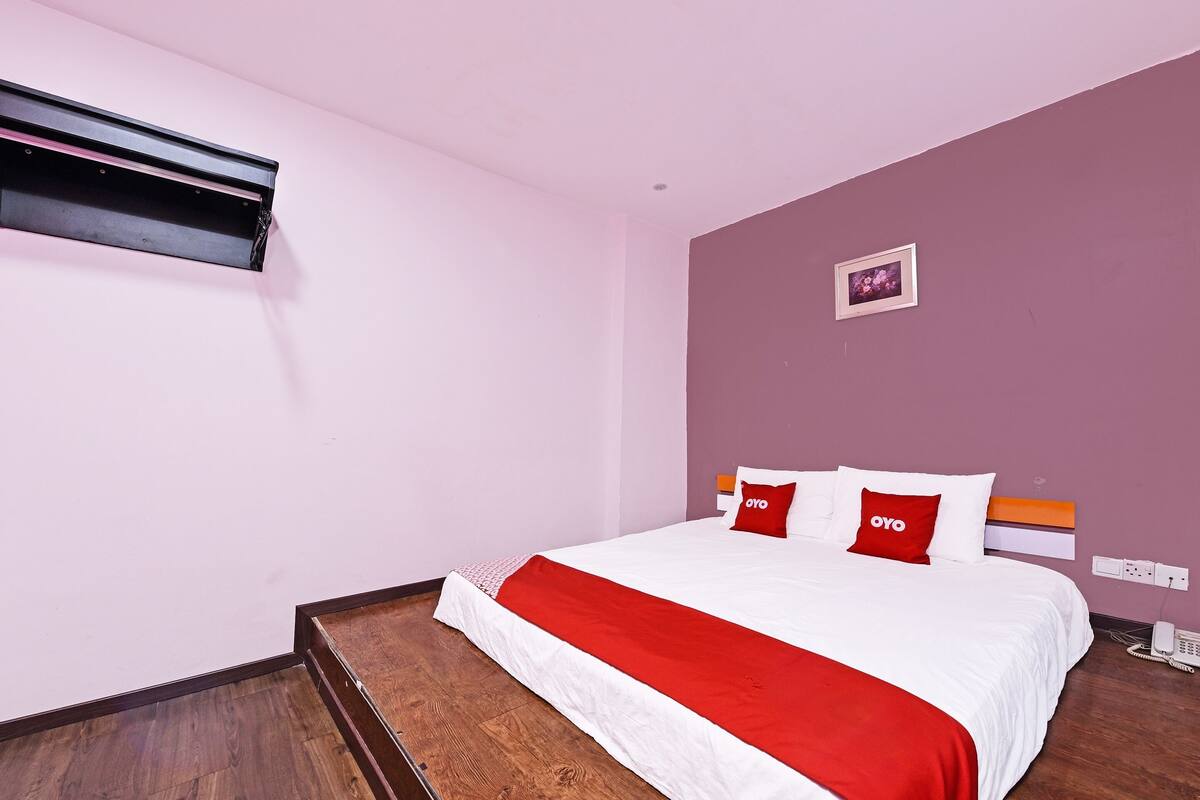 A hotel room with a bed, a red and white bedspread, a wooden floor, a wall-mounted air conditioner, and a small framed picture on the wall.