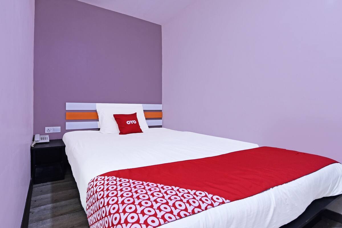 A neatly made bed with a red and white patterned bedspread, a white pillow with a red logo, and a white headboard.