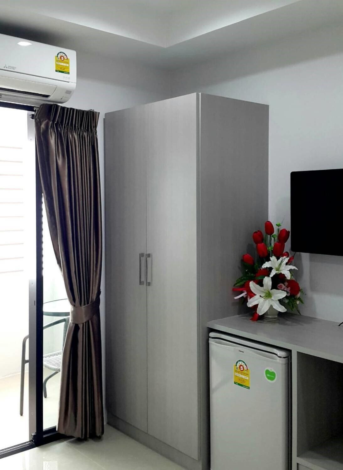 A modern hotel room with a sliding glass door, a grey wardrobe, a small fridge, and a bouquet of red and white flowers on a table.