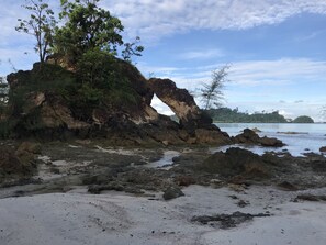 A rocky beach with a large arch formation, sandy foreground, and distant greenery.