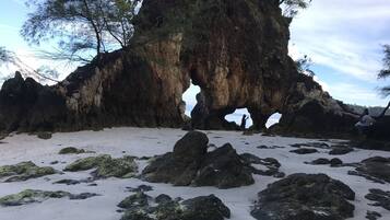 A natural rock formation with a cave-like opening, surrounded by sandy beach and scattered rocks.