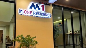 A M-One Residence sign with a logo, a potted plant, and a trash bin. - M-One Residence (Hat Yai)