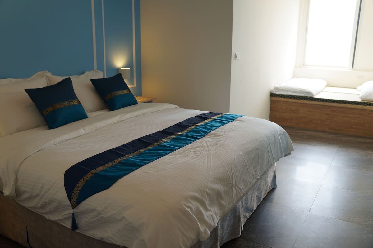 Een netjes opgemaakt bed met een blauw-witte spreideken, twee kussens en een nachtkastje met een lamp.