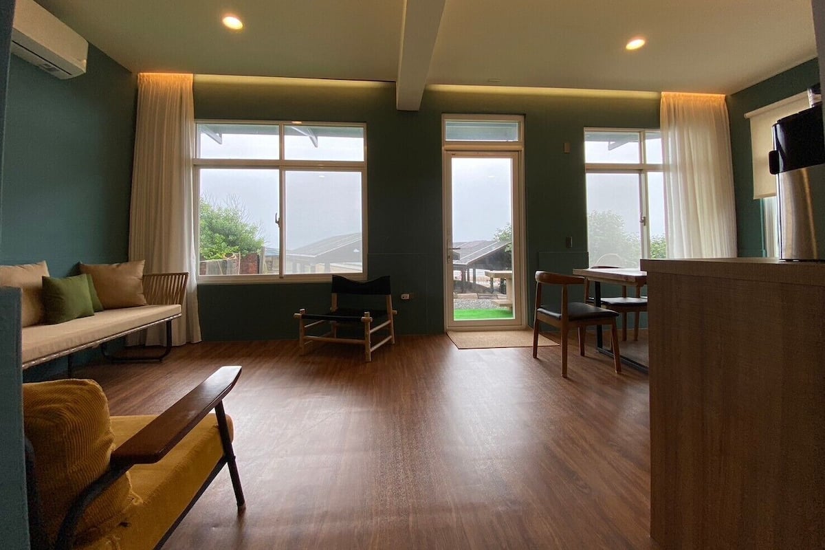 Een ruime kamer met houten vloer, een bank, een eettafel met stoelen en grote ramen voorzien van gordijnen.