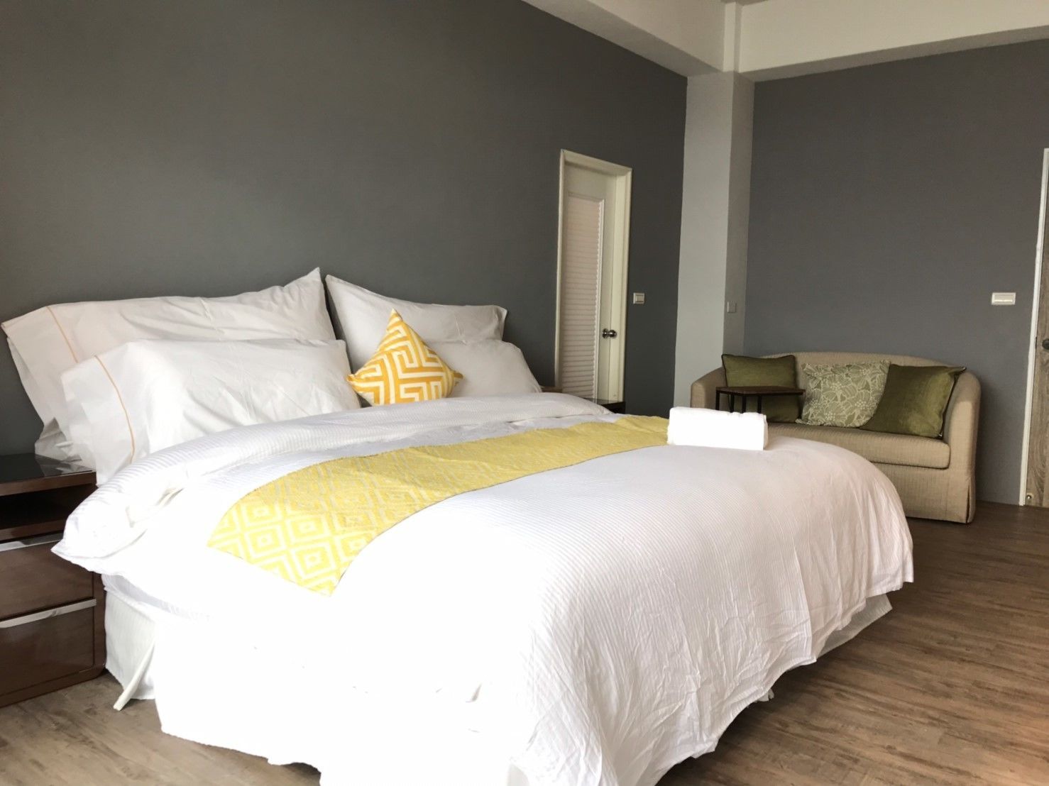 Una cama bien tendida con ropa de cama blanca y amarilla, un sofá color beige con cojines estampados, y un suelo de madera.