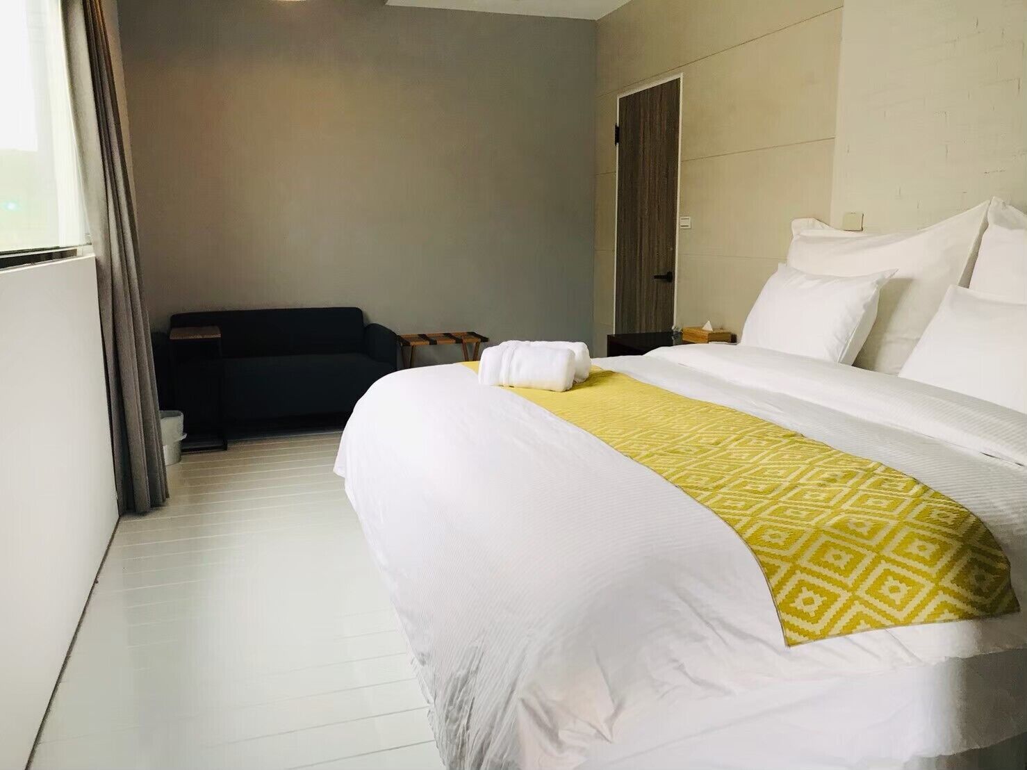 Hotellihuone, jossa on suuri sänky, tummanvärinen sohva, pieni pöytä ja ovi.