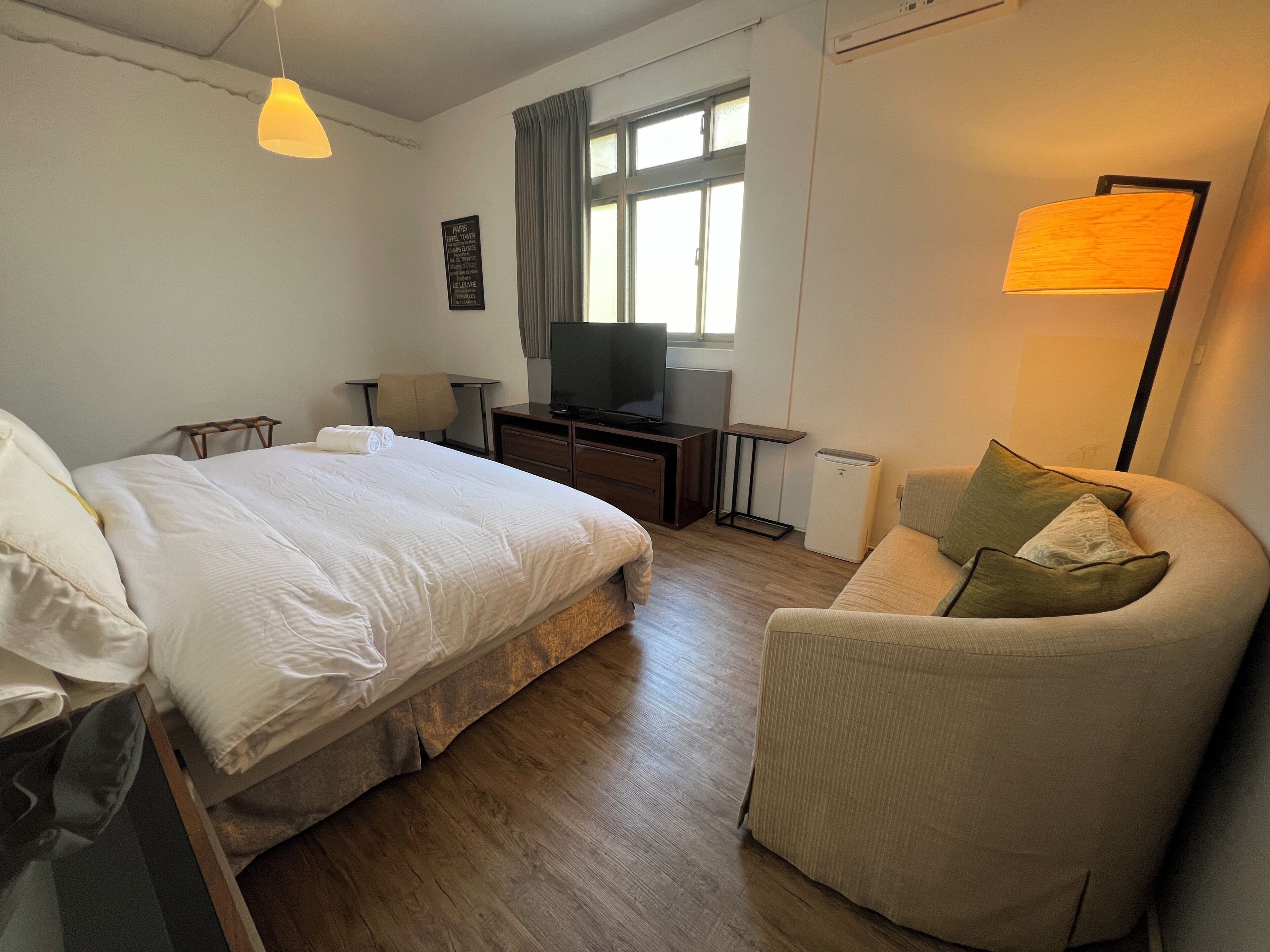 Quarto de hotel com cama, escrivaninha com computador, cadeira, sofá e televisão.