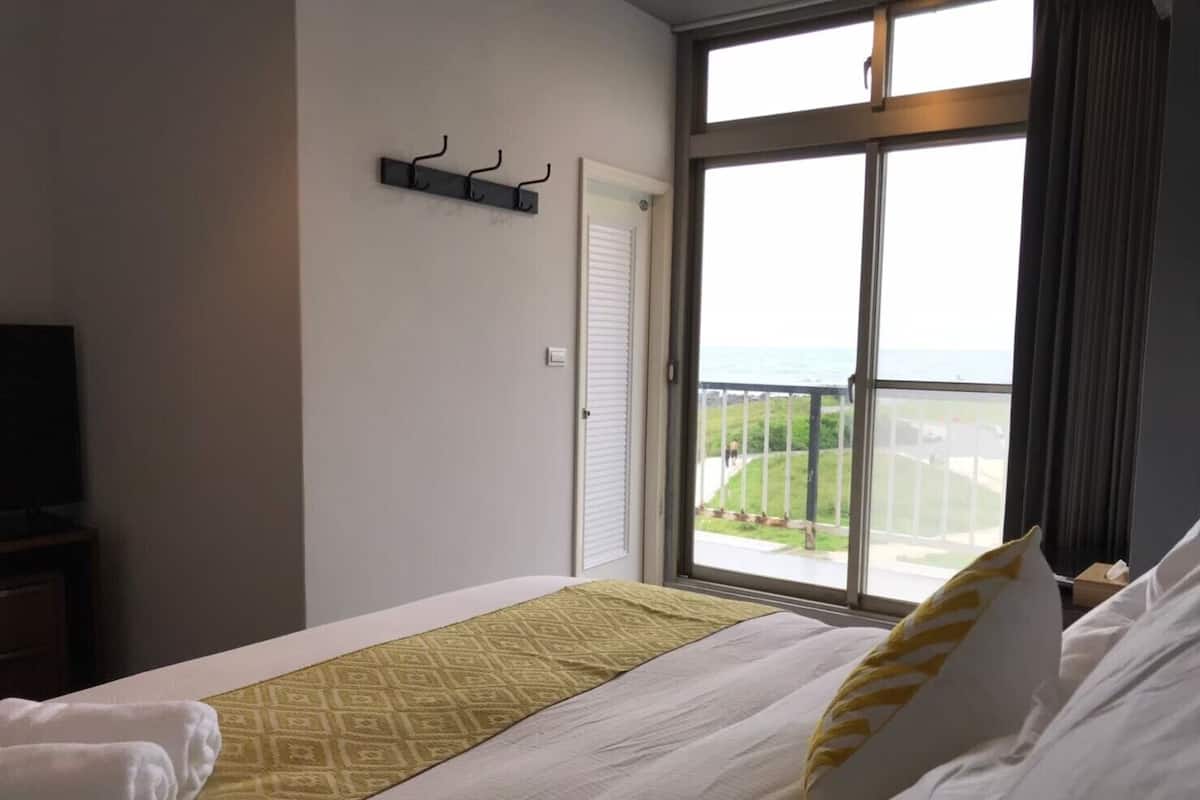 Een hotelkamer met een groot raam, een bed met wit en geel beddengoed, een televisie op een standaard en een kapstok.