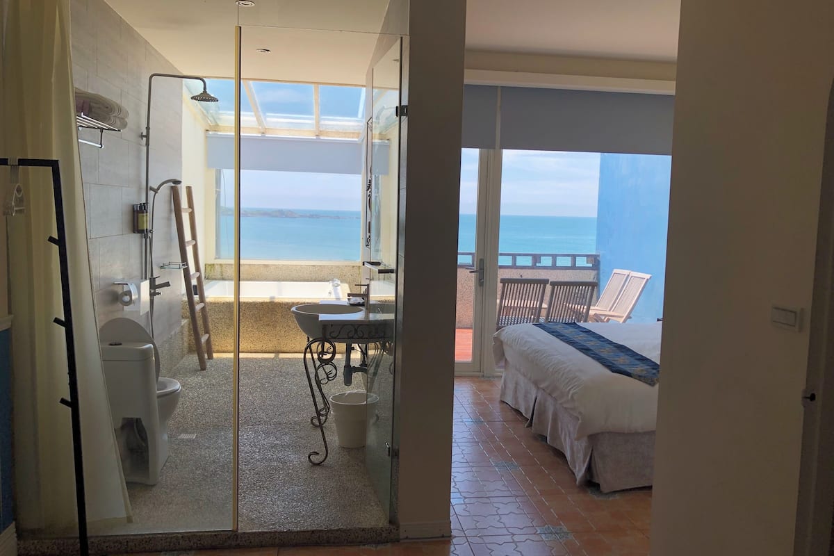Een moderne badkamer met een douchecabine van glas, een toilet en een wastafel. Er is een balkon met uitzicht op zee en een bed met uitzicht op zee.