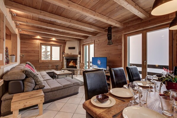 A cozy cabin interior with a fireplace, a television, a dining area with chairs and a table set for four, and a large window overlooking a snowy landscape.