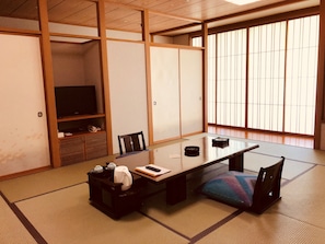 A traditional Japanese room with tatami flooring, a low table with cushions, a television on a shelf, and sliding doors. - Hotel Ichiyotei (Minakami)