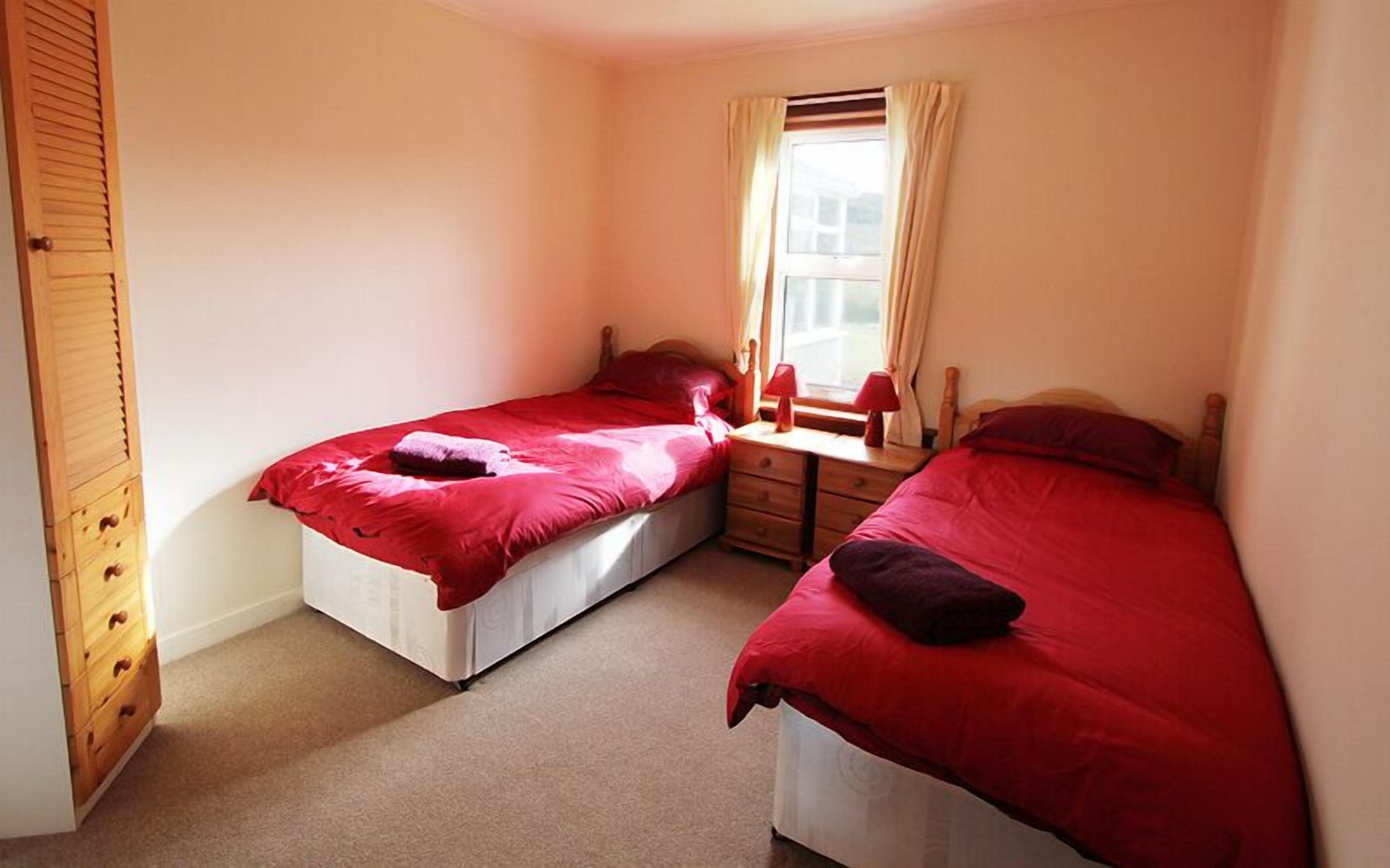 A bedroom with two single beds, a wooden wardrobe, a desk with a chair, and a window with curtains.