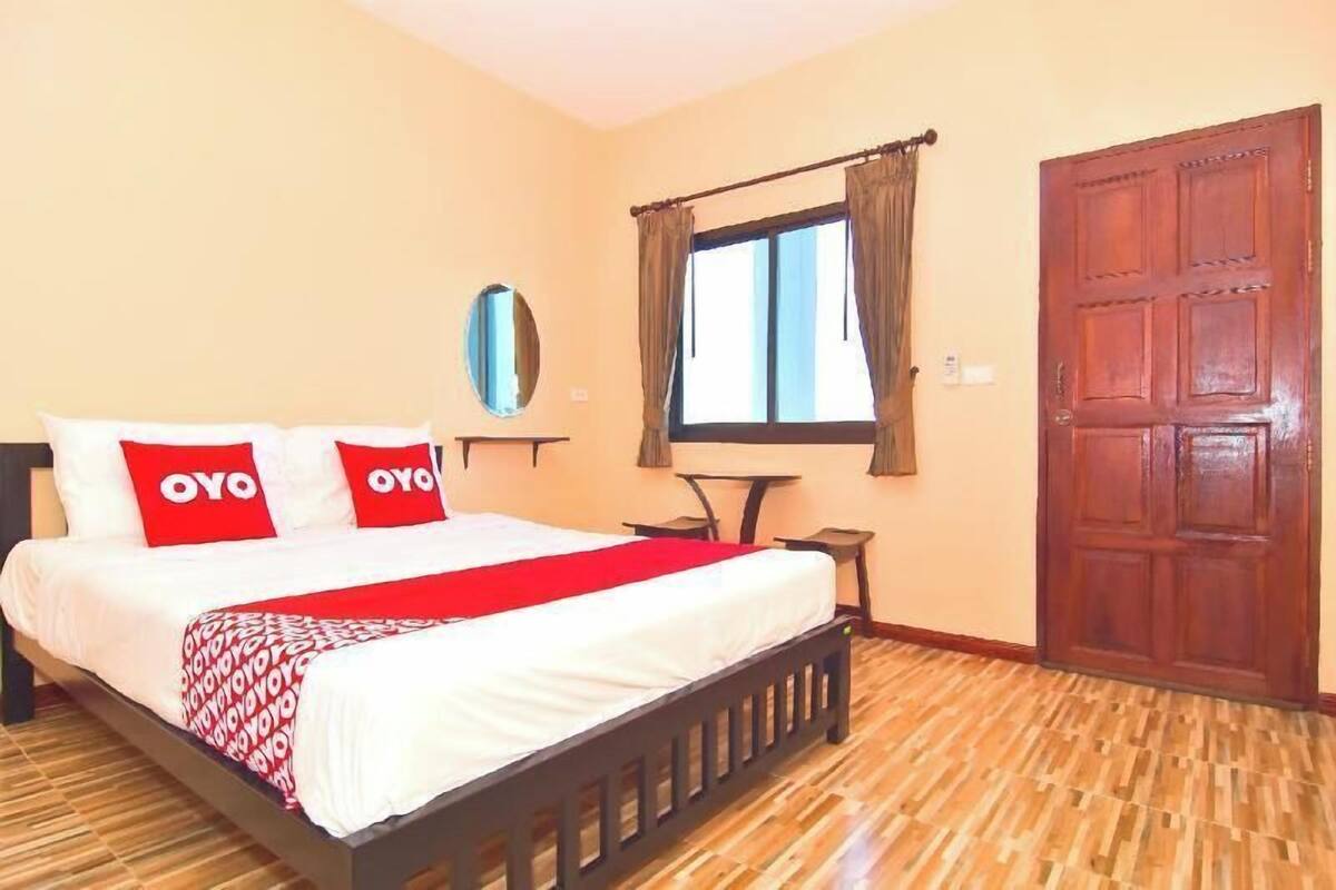 A bedroom with a bed, two pillows with 'OYO' branding, a window with curtains, a wooden door, and a small table.