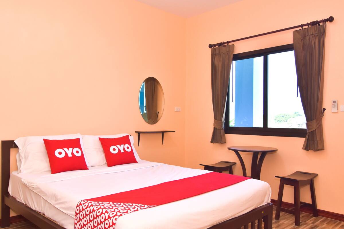 A hotel room with a bed, two red pillows, a red and white blanket, a round mirror, a window with curtains, and two small tables.