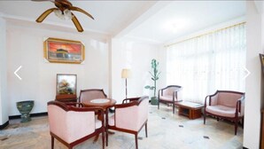Un salón amplio con techo alto, ventilador de techo, una mesa redonda para café y dos sillones. Hay una planta en maceta y un cuadro en la pared.
