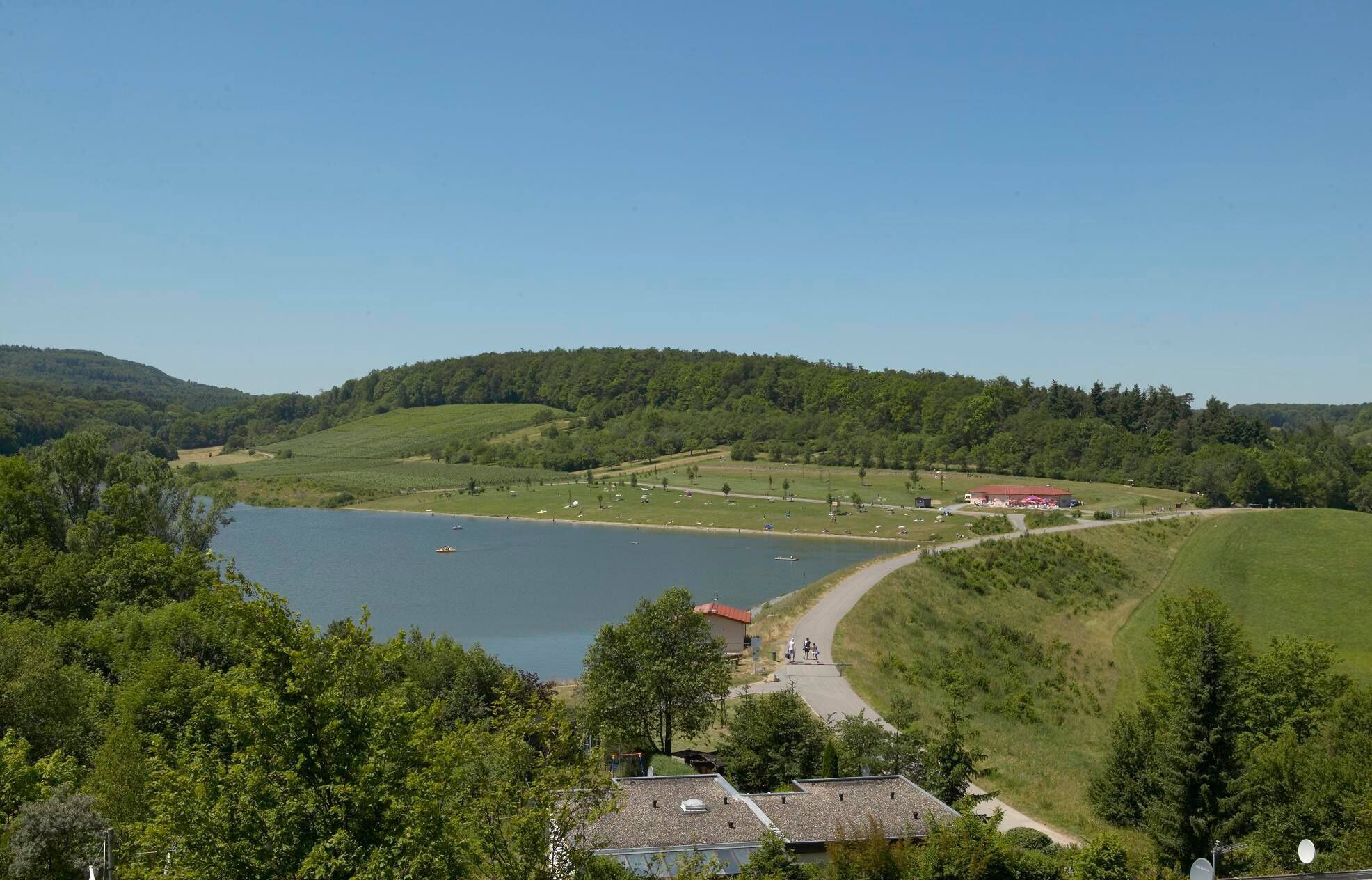 Vista d'un llac amb un camí que condueix a un edifici de teulada vermella, envoltat de verdura i turons.